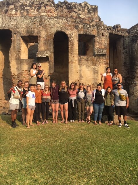 A group of students posing in front of ancient stone ruins on a grassy area. Some are standing on elevated spots.
